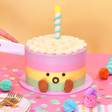 Jellycat Rainbow Birthday Cake with pastel ombre icing, decorative candle, and cute face on festive pink table by Crumbs & Doilies